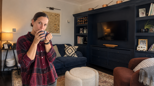 Interior designer Elise Ashley holding a mug in a warm family living room with navy built-in cabinetry and layered Modern Heritage decor.